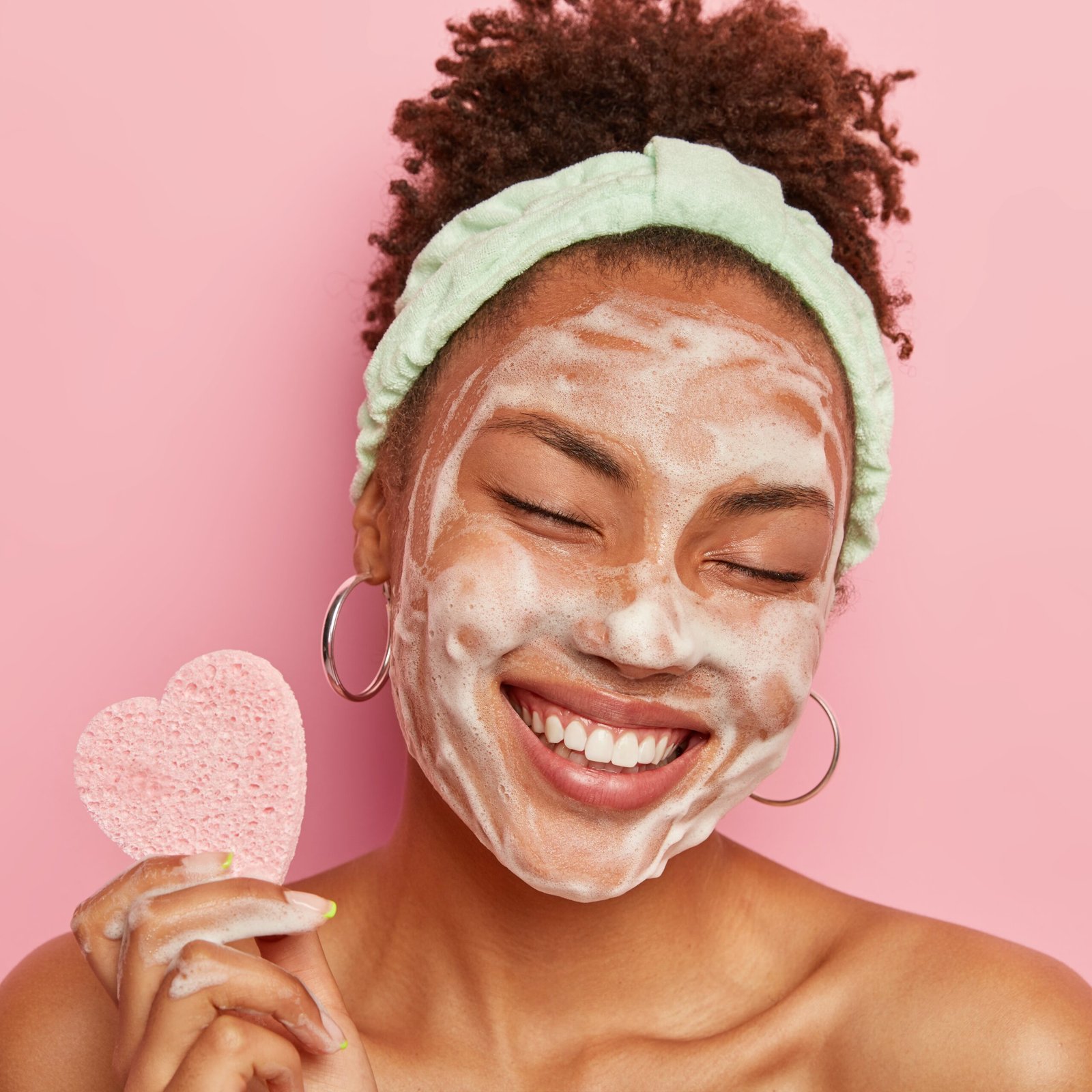 Beautiful optimistic Afro American woman cleanses face with foam, refreshes skin, has well cared complexion, holds heart shaped sponge for beauty procedures, stands bare shoulders with closed eyes